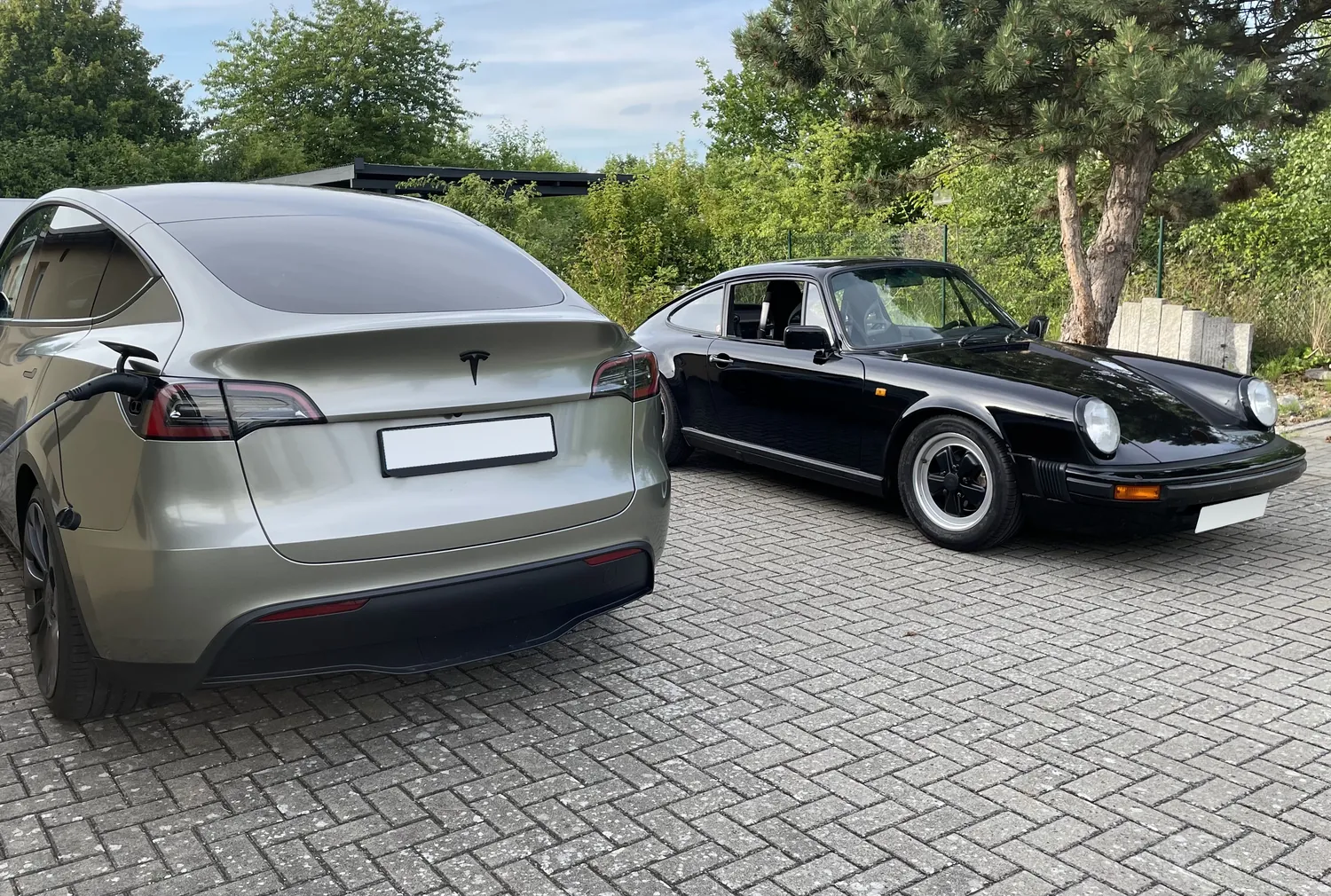 Silbernes Elektroauto am Ladekabel neben einem schwarzen Sportwagen-Klassiker